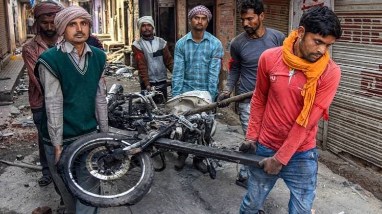 दिल्ली हिंसा में जलाई गई बाइक को लेकर जाते मजदूर (फोटो- पीटीआई)
