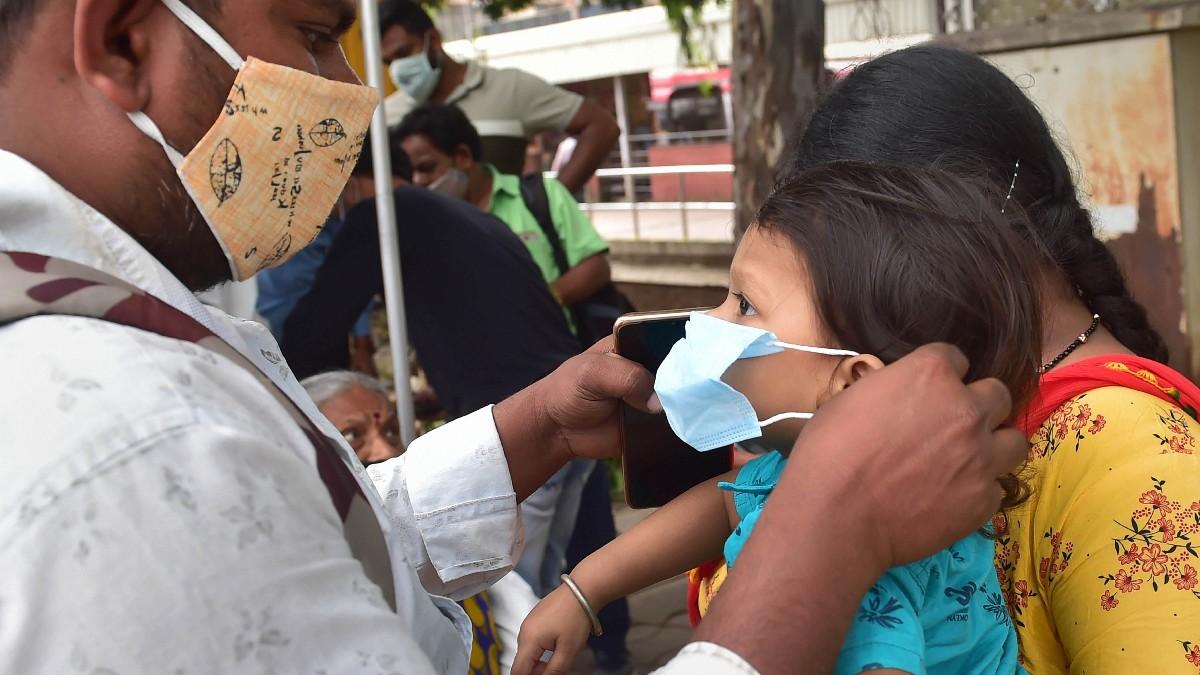 बेंगलुरु में एक बस स्टैंड पर अपने बच्चे को मास्क पहनाता पिता (फोटो- पीटीआई)