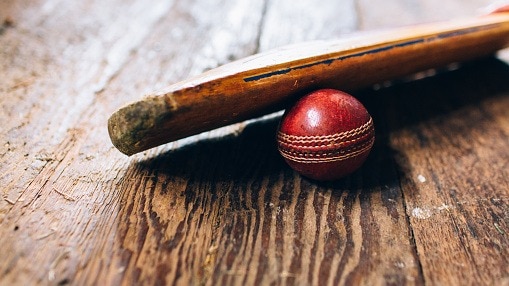 A cricket bat resting on a red cricket ball on a wooden floor in a old cricket pavilion