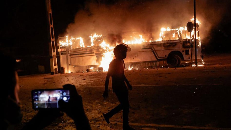 A person takes a photo as a demonstrator walks near a bus that was set on fire at the top of the road to Sri Lankan President Gotabaya Rajapaksa's residence during a protest against him as many parts of the crisis-hit country faced up to 13 hours A person takes a photo as a demonstrator walks near a bus that was set on fire at the top of the road to Sri Lankan President Gotabaya Rajapaksa's residence during a protest against him as many parts of the crisis-hit country faced up to 13 hours