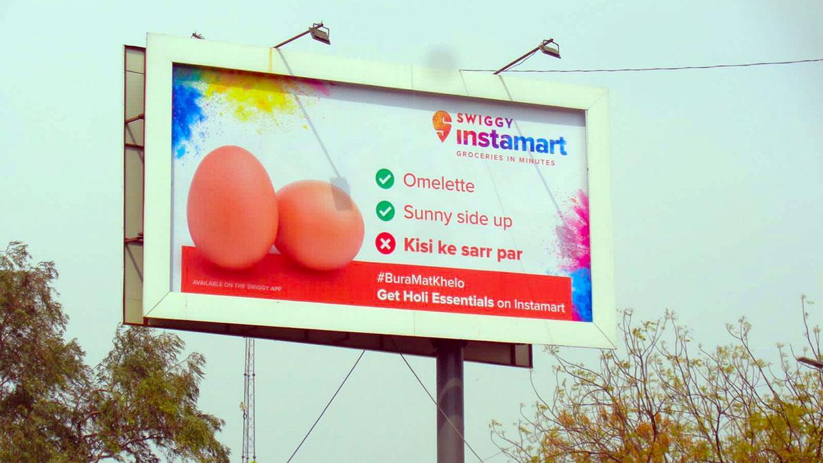 The billboard features eggs and reads, “Omelette; Sunny side-up; Kisi ke sarr par. #BuraMatKhelo. Get Holi essentials on Instamart.” The billboard features eggs and reads, “Omelette; Sunny side-up; Kisi ke sarr par. #BuraMatKhelo. Get Holi essentials on Instamart.”