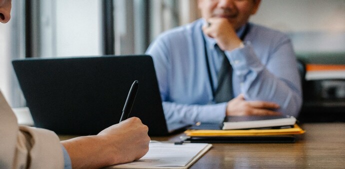 a man behind laptop on table and another writing note a man behind laptop on table and another writing note