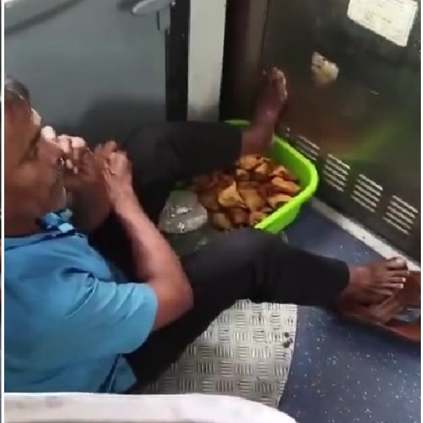 Vendor sits on floor of moving train, rests feet on basket of samosas
