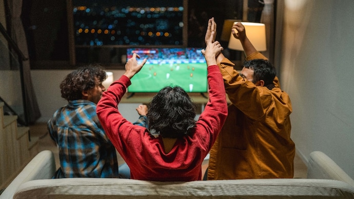 Group of excited football fans cheering and celebrating in front of the TV as their favorite team scores a goal.