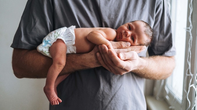 Young father with 2 weeks newborn baby boy in his hands near window at home