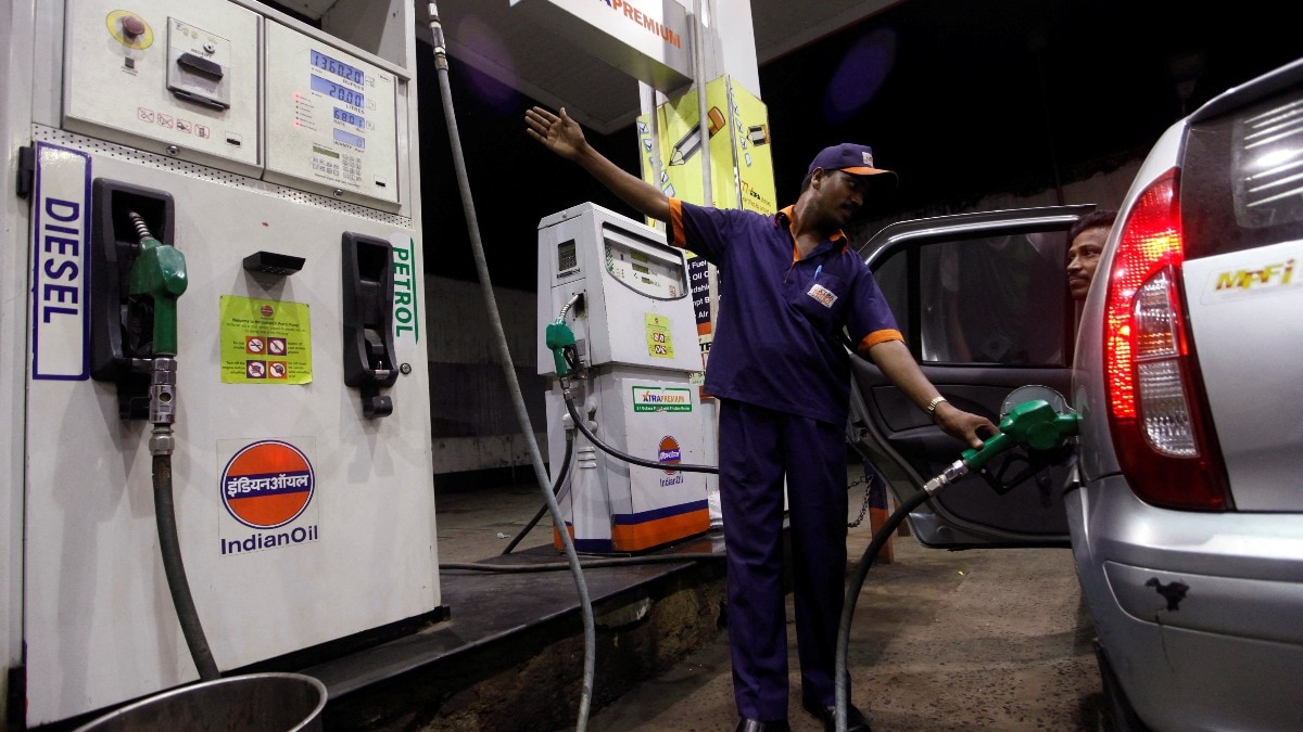 Close-up of a fuel pump nozzle dispensing E20 ethanol-blended petrol into a vehicle, with a blurred protest in the background.