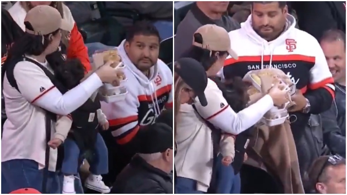 Woman carries baby, 3 plates of food as man beside her offers no help at game
