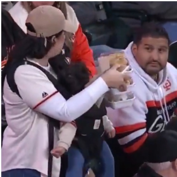 Woman carries baby, 3 plates of food as man beside her offers no help at game