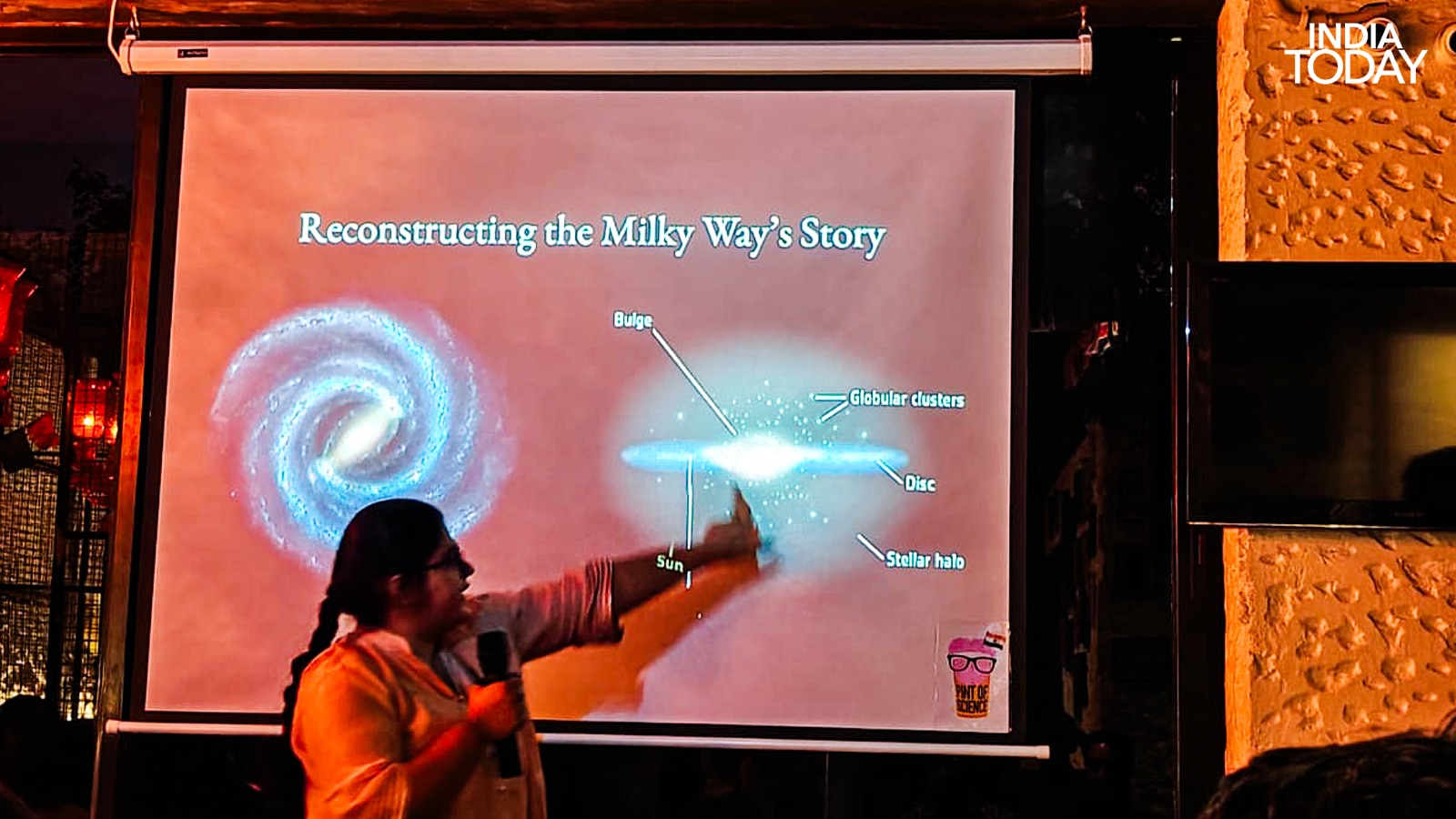 Preksha Sethia, an astrophysics scholar at University College London who studies stellar populations, reconstructing the Milky Way's story at Pint of Science 2026, Delhi. She is pointing to the position of the Sun in a galaxy that absorbed another galaxy entirely, called Gaia-Enceladus, long before Earth existed. The stars above us, she told the room, are archives. (Photo: Radifah Kabir/India Today)