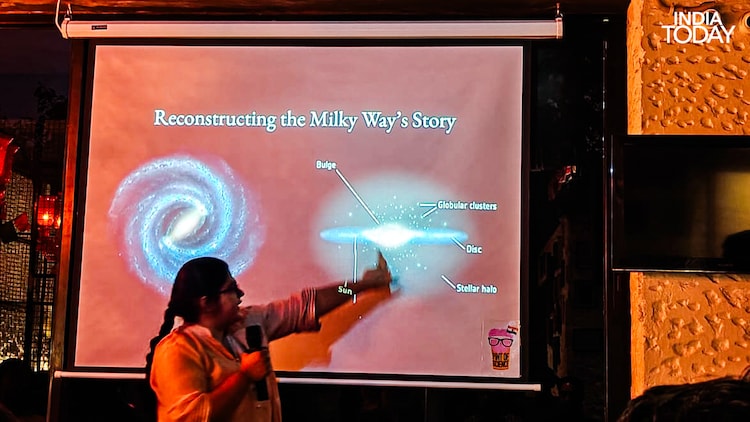 Preksha Sethia, an astrophysics scholar at University College London who studies stellar populations, reconstructing the Milky Way's story at Pint of Science 2026, Delhi. She is pointing to the position of the Sun in a galaxy that absorbed another galaxy entirely, called Gaia-Enceladus, long before Earth existed. The stars above us, she told the room, are archives. (Photo: Radifah Kabir/India Today)