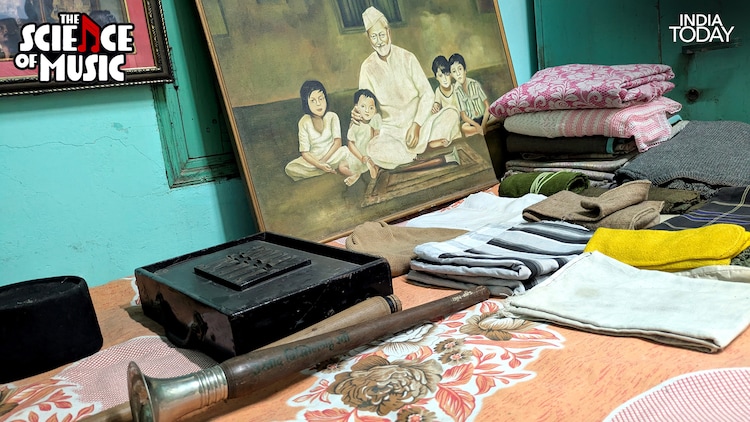 A shehnai rests in the foreground at Ustad Bismillah Khan's Varanasi home, beside a harmonium and a stack of folded clothes. The painting in the background shows the maestro seated with three children, a quiet glimpse into the life he lived between his concerts at the Red Fort and the Cannes Art Festival. (Photo: PTI)