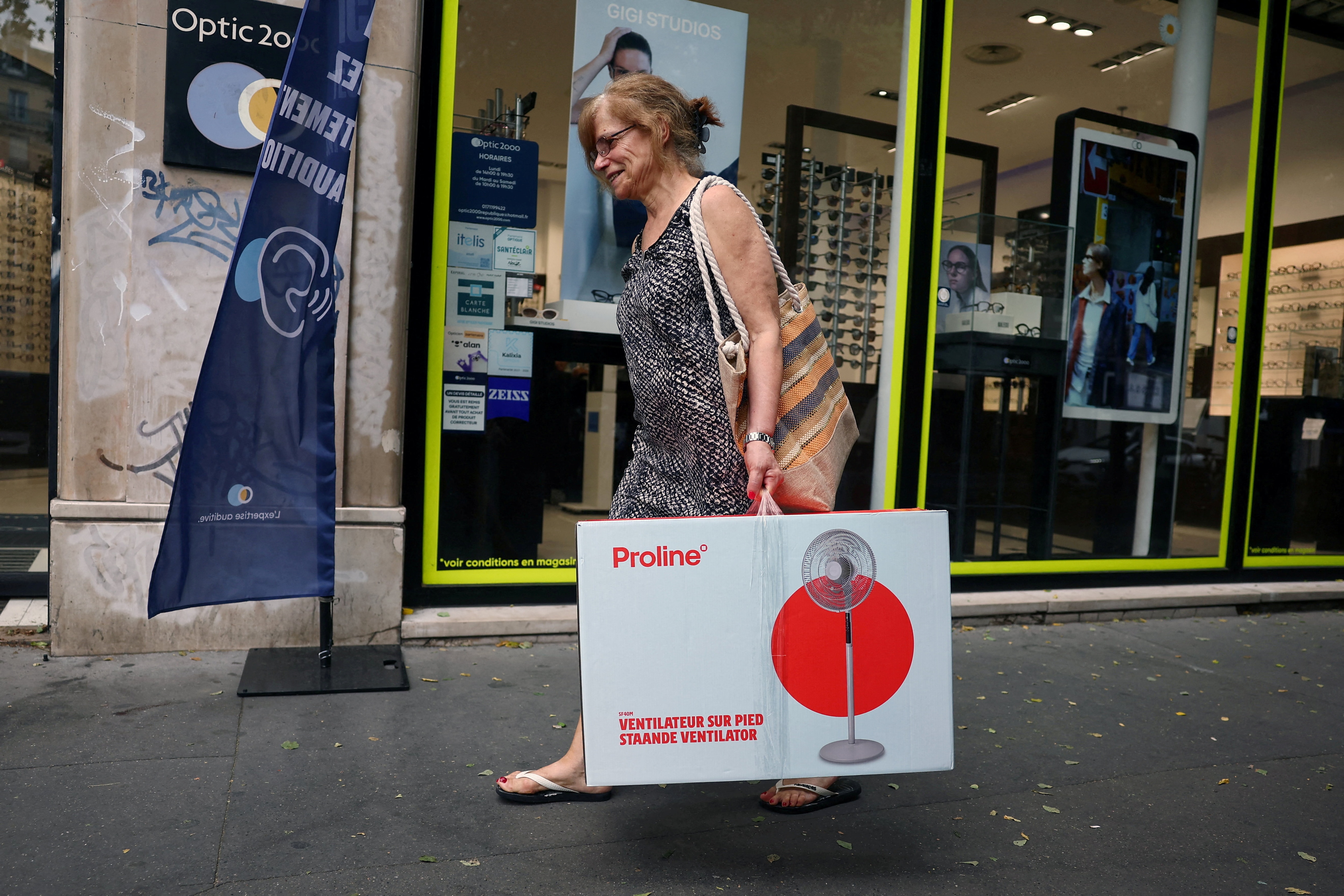 A woman carrying an electric fan in a cardboard box walks down a street 